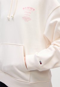 Felpa crema realizzata in morbido tessuto, con tasca frontale, branding rosa e un dettaglio del logo sul polsino con accenti rosso, bianco e blu.