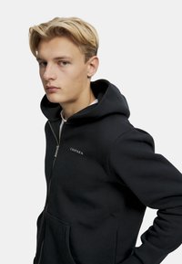 Svart zip-up hettegenser laget av mykt stoff, med kengurulomme, snorhette og subtil logo på brystet. Glatt tekstur.