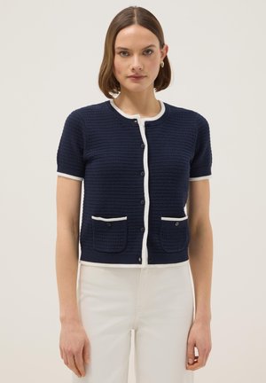 Femme portant un cardigan texturé bleu marine à manches courtes avec bordures blanches et boutons, associé à un pantalon blanc taille haute, posant devant un fond uni.