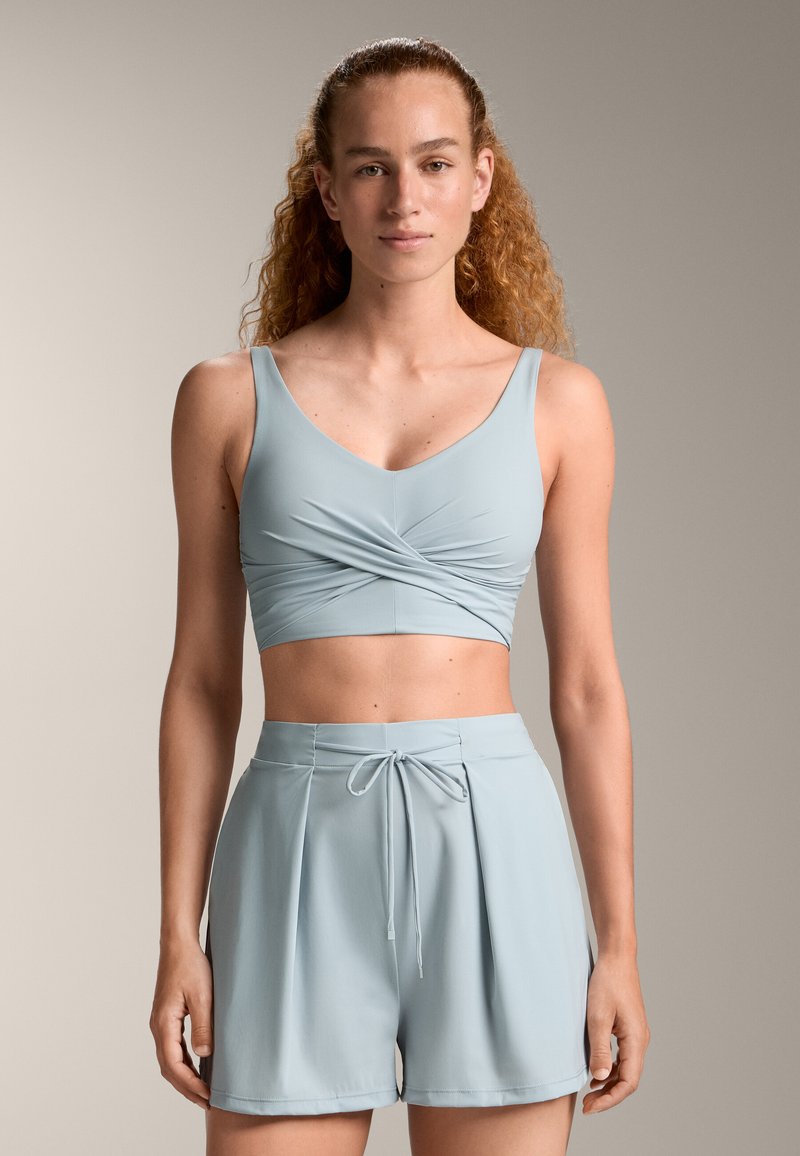 Svetlo modra aktivna obleka, ki vključuje skrajšan top z zvitim sprednjim delom in hlače s visokim pasom, ki imajo vrvico in nabran dizajn.