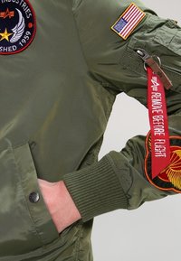 Giacca bomber verde oliva con finitura lucida, dotata di una grande toppa, bandiera americana e una etichetta rossa "Rimuovere prima del volo".