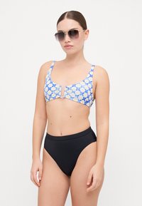 Bikiniöverdel med blå och gula blommönster, drapering i mitten, matchad med enfärgade svarta högmidjade underdelar. Modell som bär solglasögon.