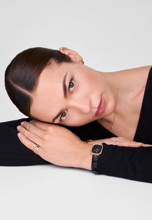 Montre en cuir noir avec un cadran rectangulaire doré et noir, accompagnée de bagues dorées superposées sur les doigts et d'un collier délicat en or.