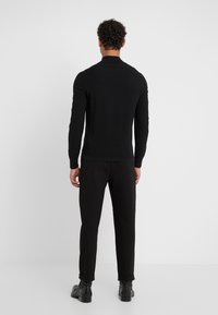 Homme aux cheveux bouclés portant une chemise noire à manches longues, un pantalon noir et des chaussures noires, debout face à un mur blanc uni.