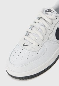 Bijeli Nike Force 1 tenisice izbliza prikazuju crni Swoosh, bijele vezice, perforirani nos i teksturiranu rubnu podlogu na svijetloj pozadini.