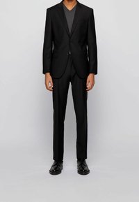 Schwarzer Anzug mit einem maßgeschneiderten Blazer mit zwei Knöpfen und passenden Hosen. Getragen über einem dunkelgrauen V-Ausschnitt-Shirt, kombiniert mit schwarzen Schuhen.