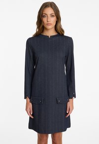 Navyblaues gestreiftes Kleid mit hohem Ausschnitt, langen Ärmeln und zwei Fronttaschen mit Knopfakzenten. Hergestellt aus strukturiertem Stoff.