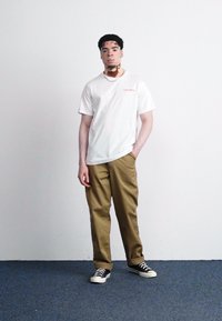 White cotton t-shirt with red text, beige trousers, and black sneakers. Simple design with a relaxed fit and rolled-up pant cuffs.