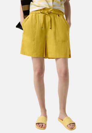 Persoon draagt gele shorts met trekkoord en zijzakken, en bijpassende gele slippers, staat met handen in de zakken.