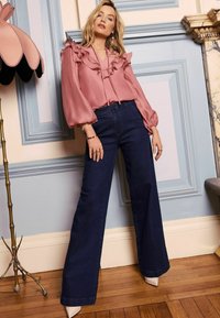 Poros rózsaszín blúz fodros részletekkel, ellensúlyozva széles szárú sötét farmernadrággal. A modell hegyes bézs magassarkút visel, és kék panelek előtt áll.