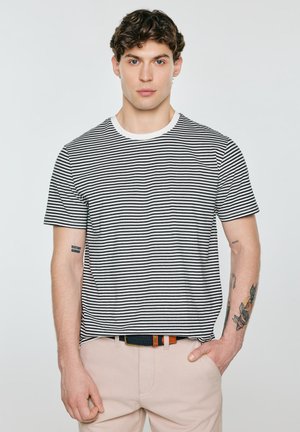 Młody mężczyzna z krótkimi kręconymi włosami, ubrany w czarno-biały paskowany t-shirt i beżowe spodnie, stoi z jedną ręką w kieszeni, ma tatuaże na ramionach.