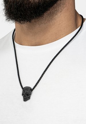 Black skull pendant necklace on a braided black cord, featuring intricate detailing on the skull surface, worn over a white shirt.