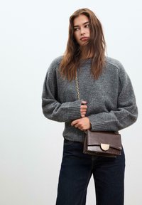 Grauer gestrickter Pullover mit gerippten Bündchen, kombiniert mit dunklen Jeans. Eine kleine braune Leder-Umhängetasche hat einen metallischen Verschluss.