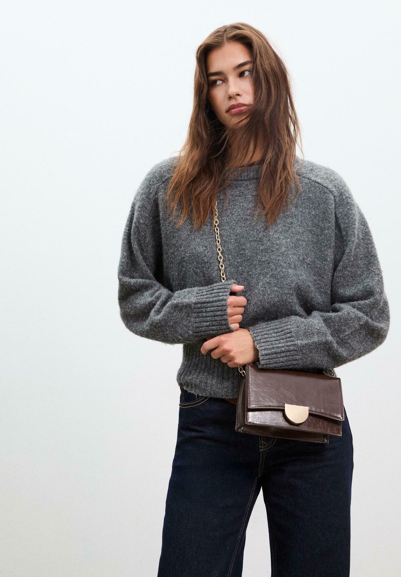 Grauer gestrickter Pullover mit gerippten Bündchen, kombiniert mit dunklen Jeans. Eine kleine braune Leder-Umhängetasche hat einen metallischen Verschluss.