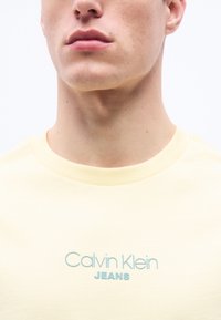 Κοντινό πλάνο του κάτω μέρους του προσώπου και του λαιμού ενός ατόμου που φορά ένα ανοιχτό κίτρινο t-shirt με την επιγραφή "Calvin Klein JEANS" σε ανοιχτό μπλε χρώμα.
