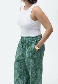 Femme portant un haut blanc sans manches et un pantalon vert taille haute à motif cachemire, les mains dans les poches et un bracelet en or.
