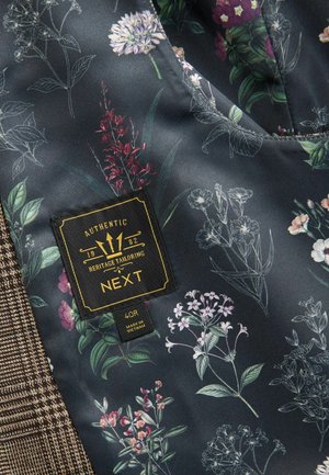 Stoff mit floralen Mustern in Dunkelblau mit grünen Akzenten, ausgestattet mit einer gewebten, hellbraunen Textur und einem schwarzen Etikett mit goldener Schrift.