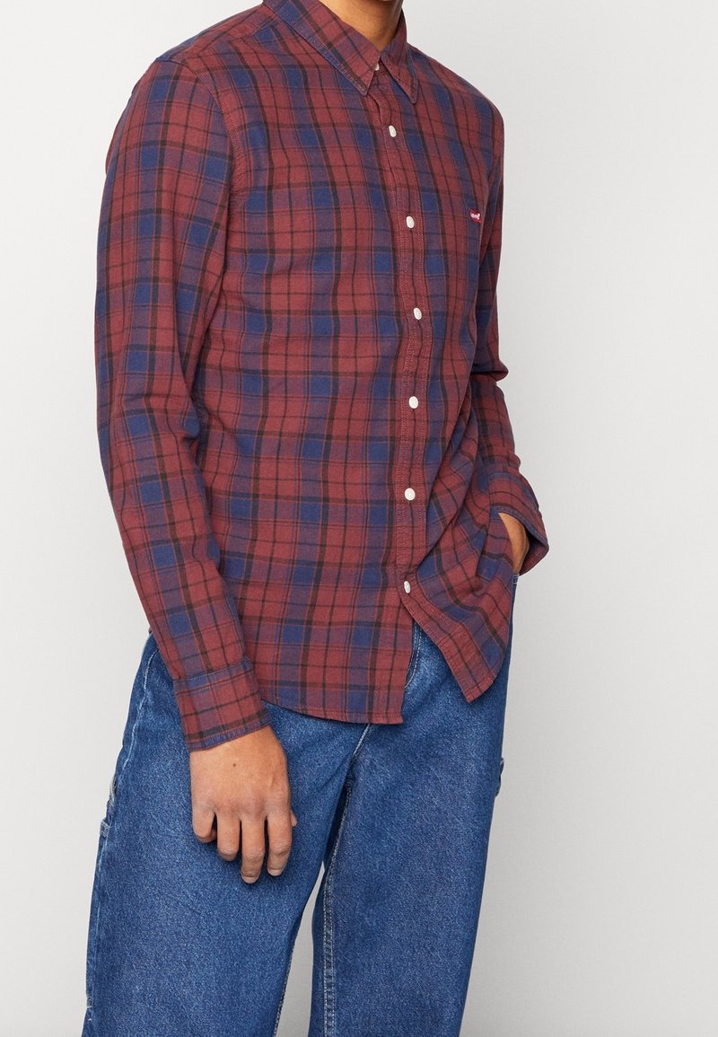 Homme portant une chemise à carreaux rouge et bleue à manches longues avec boutons et un jean bleu en denim, une main dans la poche.