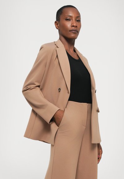 Beige skreddersydd blazer med spiss krage, dobbeltspent design og lommer, kombinert med en svart tettsittende topp og matchende bukser.