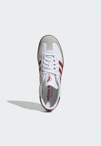 Basket en cuir blanc Adidas avec des rayures rouges et un bout en daim beige, vue d'en haut, projetant une ombre sur un fond blanc.