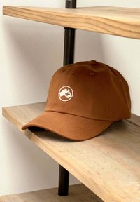 Gorra marrón hecha de tela texturizada, con un logo blanco circular que presenta un diseño. La visera curvada descansa sobre una estantería de madera.