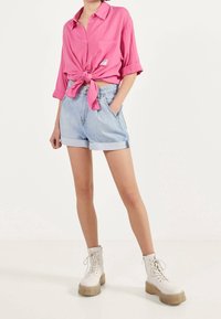 Osoba w jasnoróżowej koszuli zapinanej na guziki związanej w talii, błękitnych, podwiniętych szortach dżinsowych i białych, masywnych butach na sznurówki.
