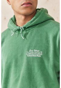 Zelená mikina s kapucí z měkké látky, s kapucí na stažení, bílým nápisem na přední straně a viditelnými švy.