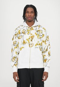 White zip-up hoodie featuring a yellow and black swirling floral pattern, cotton material, and front pockets. Long sleeves with ribbed cuffs.
