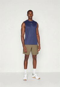 Muž stojí čelom dopredu, oblečený v námorníckom bezrukávovom športovom tričku, olivových šortkách, bielych ponožkách a bielych športových topánkach pred jednoliatym pozadím.