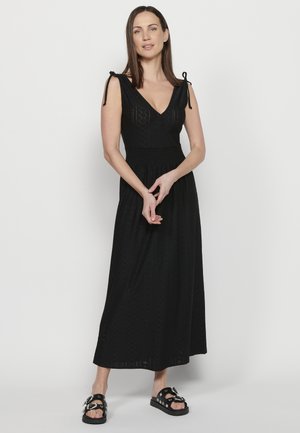 Abito lungo nero con scollatura a V, spalline regolabili con lacci e vita arricciata. Presenta un tessuto testurizzato con un design a motivi. Sandali neri con decorazioni.