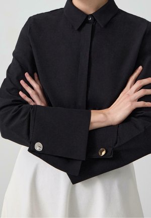 Persoon met gekruiste armen die een zwarte lange mouwen blouse draagt met niet-overeenkomende manchetknopen en een witte rok of broek.