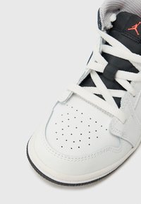 Λευκό δερμάτινο αθλητικό παπούτσι με μαύρες λεπτομέρειες. Διαθέτει διάτρητο μπροστινό μέρος, παχιά λευκά κορδόνια και χαρακτηριστικό λογότυπο σε πορτοκαλί. Φαίνεται η υφασμένη σόλα.