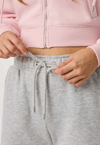 Person, der justerer snoren på lysegrå joggingbukser, iført en pink lynlåstrøje, der viser maven.