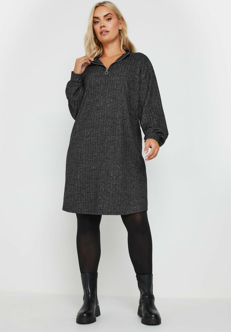 Abito nero a coste con colletto a mezza zip, maniche oversize e vestibilità rilassata, abbinato a collant neri e stivali neri robusti.