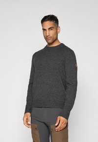 Mand iført en mørkegrå strikket sweater og mørke bukser med brune cargo-lommer, som står foran en ensfarvet lys baggrund.