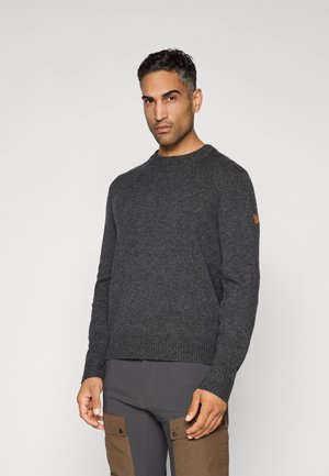 Man in een donkergrijs gebreid vest en donkere broek met brown cargozakken, staande tegen een effen lichte achtergrond.