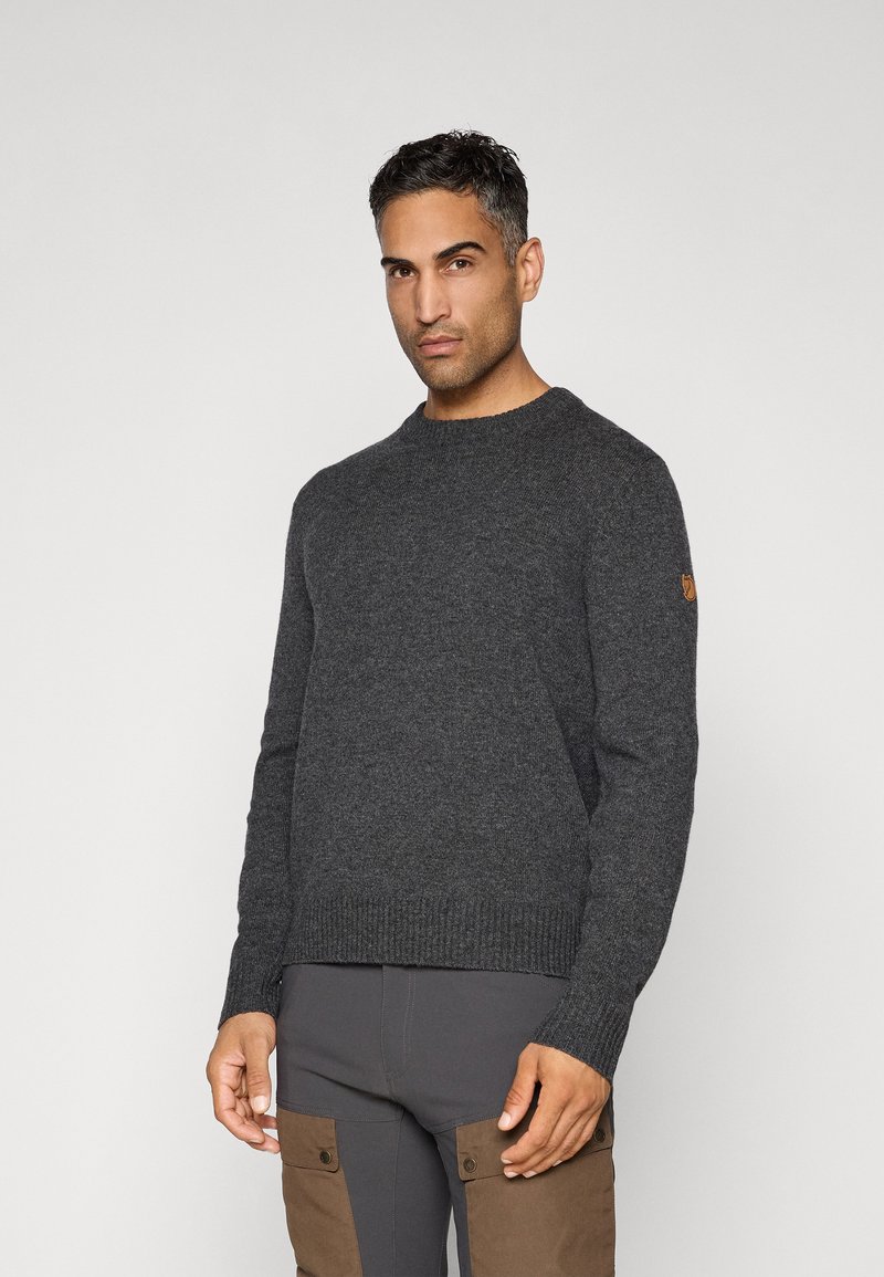 Mand iført en mørkegrå strikket sweater og mørke bukser med brune cargo-lommer, som står foran en ensfarvet lys baggrund.