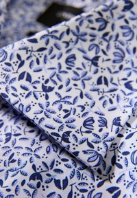 Witte stof met een gedetailleerd blauw bloemmotief, met bladeren en bloemen. De kraag heeft een scherpe, gestructureerde vorm.