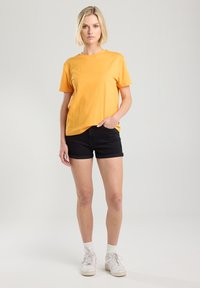 T-shirt en coton jaune avec une coupe décontractée, associé à des shorts en denim noirs. Le modèle porte des baskets blanches et a une coiffure décontractée.