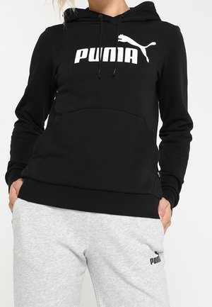 Personne portant un sweat à capuche Puma noir et un pantalon de survêtement Puma gris clair, les mains dans les poches, debout devant un fond uni.