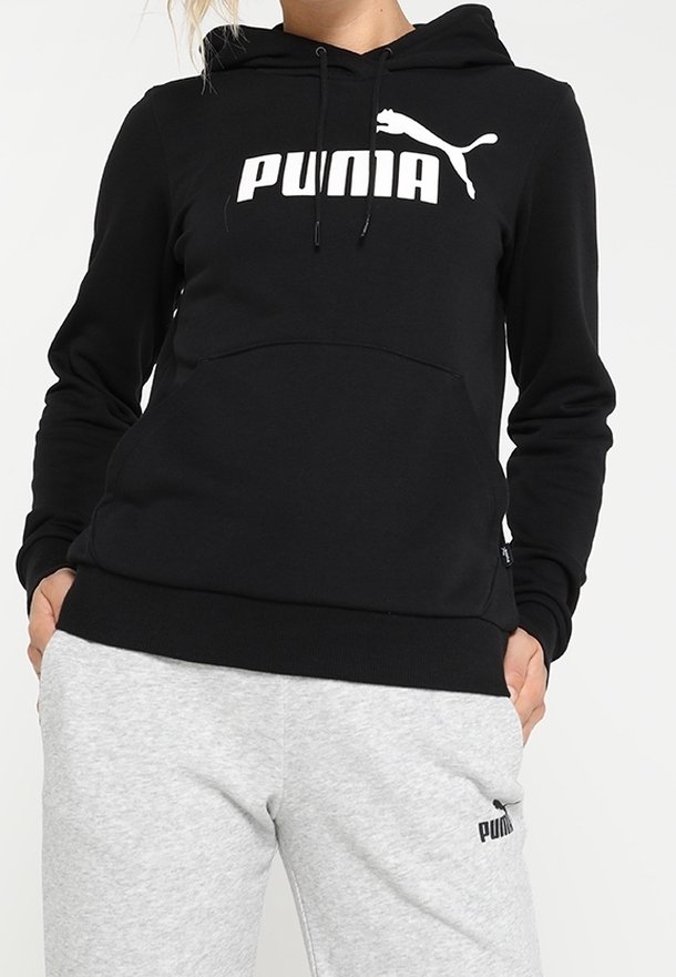 Personne portant un sweat à capuche Puma noir et un pantalon de survêtement Puma gris clair, les mains dans les poches, debout devant un fond uni.