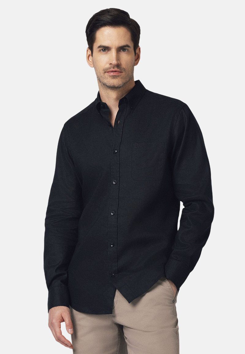Czarna koszula zapinana na guziki z kołnierzykiem, długimi rękawami i przednią kieszenią. Wykonana z teksturowanej tkaniny, noszona z jasnymi spodniami.