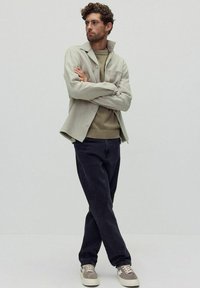 Ljusgrå skjorta över en beige tröja, tillsammans med mörka jeans och beige sneakers. Modellen står med armarna i kors mot en enkel bakgrund.