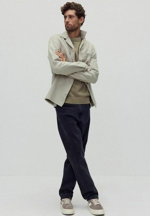 Lichtgrijs overhemd met knopen over een beige trui, gecombineerd met donkere jeans en beige sneakers. Model staat met gekruiste armen tegen een effen achtergrond.