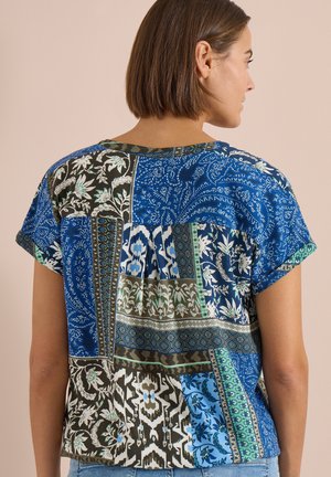 Kvinde med kort brunt hår iført en blå og grøn patchwork-blomstret kortærmet bluse, set bagfra.