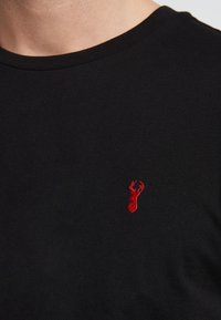 Musta puuvillainen T-paita, jossa on pyöreä pääntie ja pieni punainen kirjailtu hummerilogo vasemmalla puolella rinnassa.