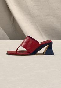 Κόκκινο δερμάτινο mule πέδιλο με ανοιχτά δάχτυλα, φαρδιά λωρίδα, θηλιά για τα δάχτυλα και σκαλιστή μαύρη φιάπα τακουνιού πάνω σε ανοιχτή επιφάνεια.