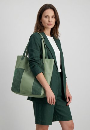 Frau in dunkelgrünem Blazer und Shorts trägt eine große zweifarbige grüne Ledertasche über der Schulter.