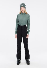 Haut à col roulé côtelé vert, pantalon de ski noir taille haute avec fermeture par bouton et zip, porté avec des bottes de ski noires et un bonnet vert assorti.