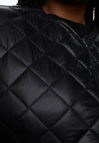 Gros plan d'une personne portant une veste matelassée noire avec des coutures en forme de losange et une fermeture éclair à l'avant.